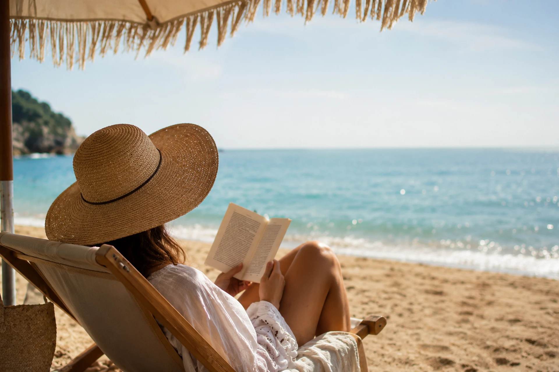 Kvinne med solhatt leser en bok i strandstol ved havet